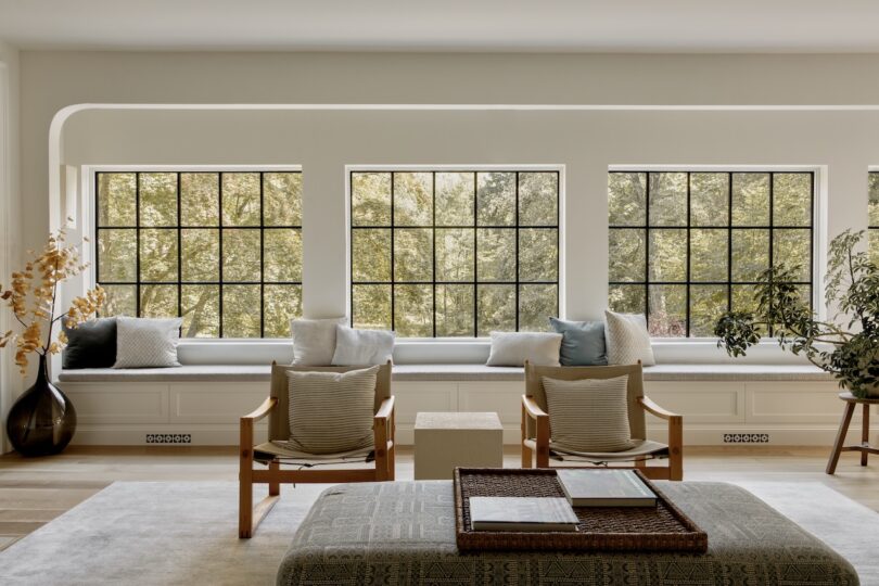 Bright living room with large grid windows, a window seat with cushions, two armchairs, a cushioned ottoman, and indoor plants. Trees are visible outside.
