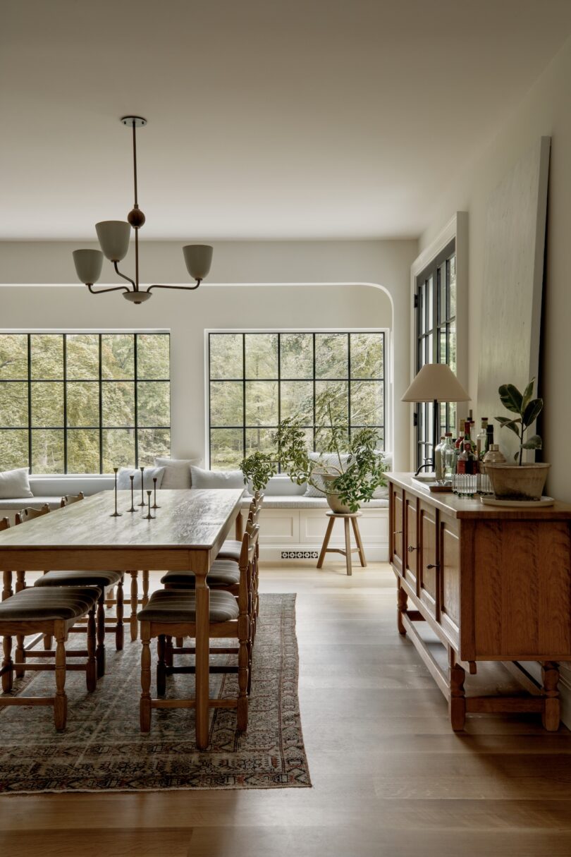 A dining room with a wooden table, chairs, sideboard, rug, potted plants, and large windows letting in natural light.