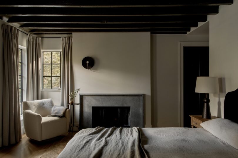 A bedroom with a bed, armchair by a window, fireplace, nightstand with lamp, and exposed wooden ceiling beams.
