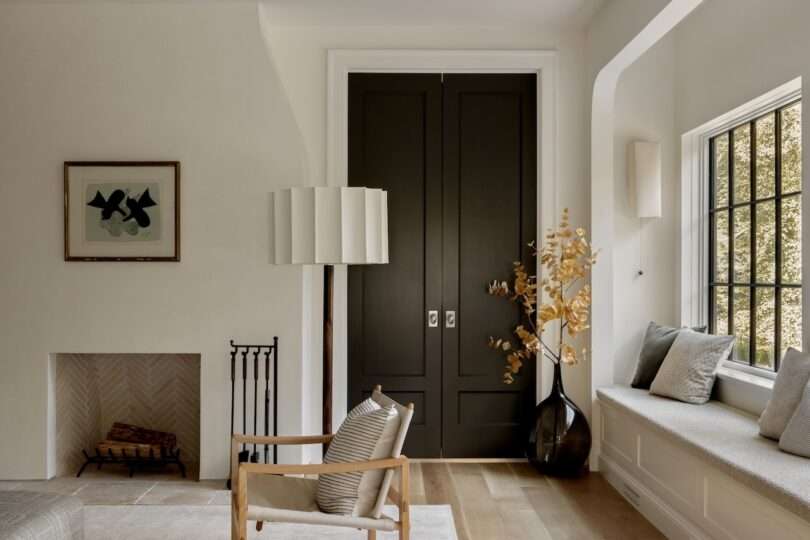 Minimalist living room with a black double door, neutral walls, a fireplace, wooden chair, floor lamp, window bench, and a vase with dried branches.