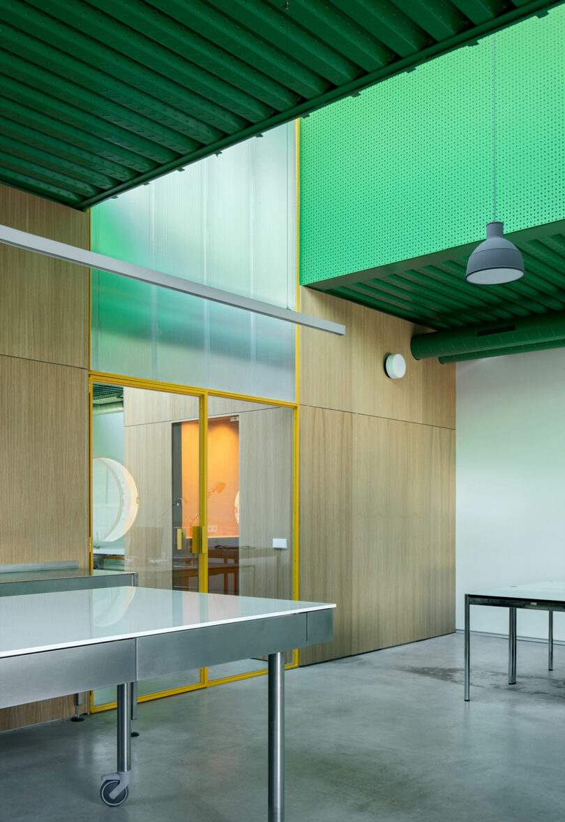 Modern interior at Impepinable Studio with green ceiling panels, wood-paneled walls, glass doors, and metal tables on a polished concrete floor. Exposed ceiling lights and ventilation pipes add an industrial touch.