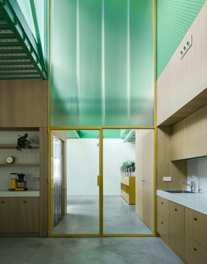 Modern kitchen by Impepinable Studio with wood cabinetry, tiled backsplash, coffee maker, and a yellow-framed glass door leading to a hallway featuring green translucent ceiling panels and concrete flooring.