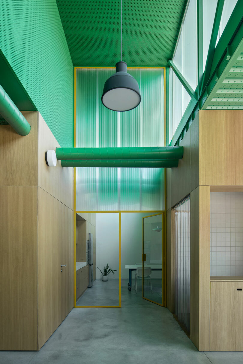 Modern interior by Impepinable Studio with green ceiling, wood panel walls, large hanging lamp, glass doors with yellow frame, and minimalist office furniture. Natural light enters from high windows.
