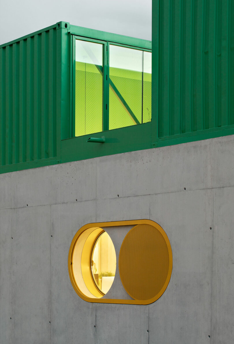 A green Impepinable Studio shipping container structure sits atop a concrete wall with a circular yellow-framed window featuring a sliding cover.
