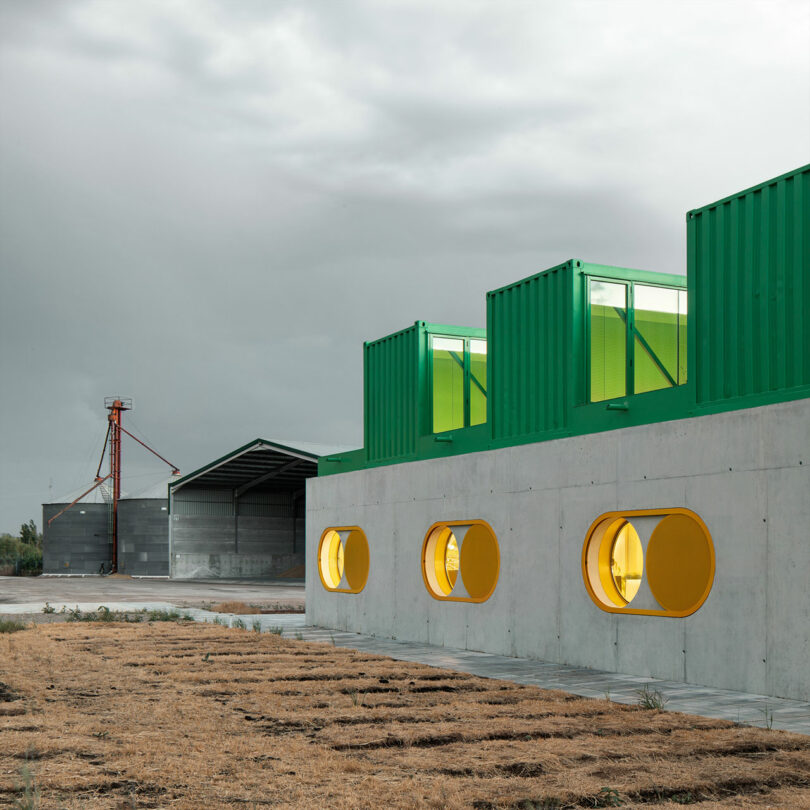 Modern industrial building, known as Impepinable Studio, with green shipping containers on the roof and round yellow windows, adjacent to a field and another large shed under a cloudy sky.