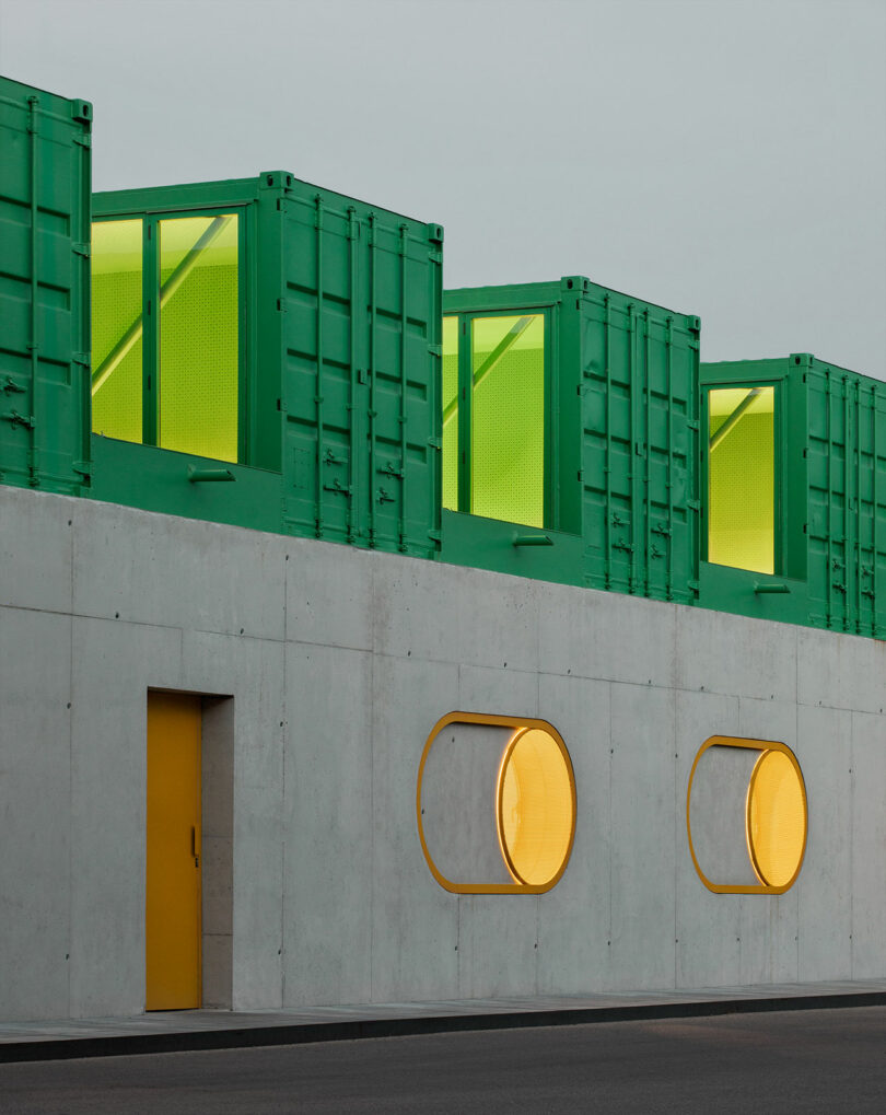 A modern building with a gray concrete facade, round yellow windows, a yellow door, and stacked green shipping containers on the roof—this is the signature look of Impepinable Studio.