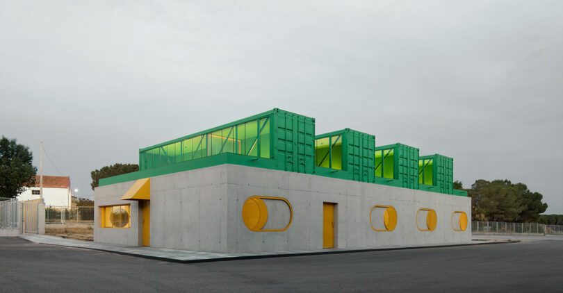 A modern Impepinable Studio building with a gray concrete base, green structures on the roof, and yellow circular windows and doors, situated on a paved lot with trees in the background.