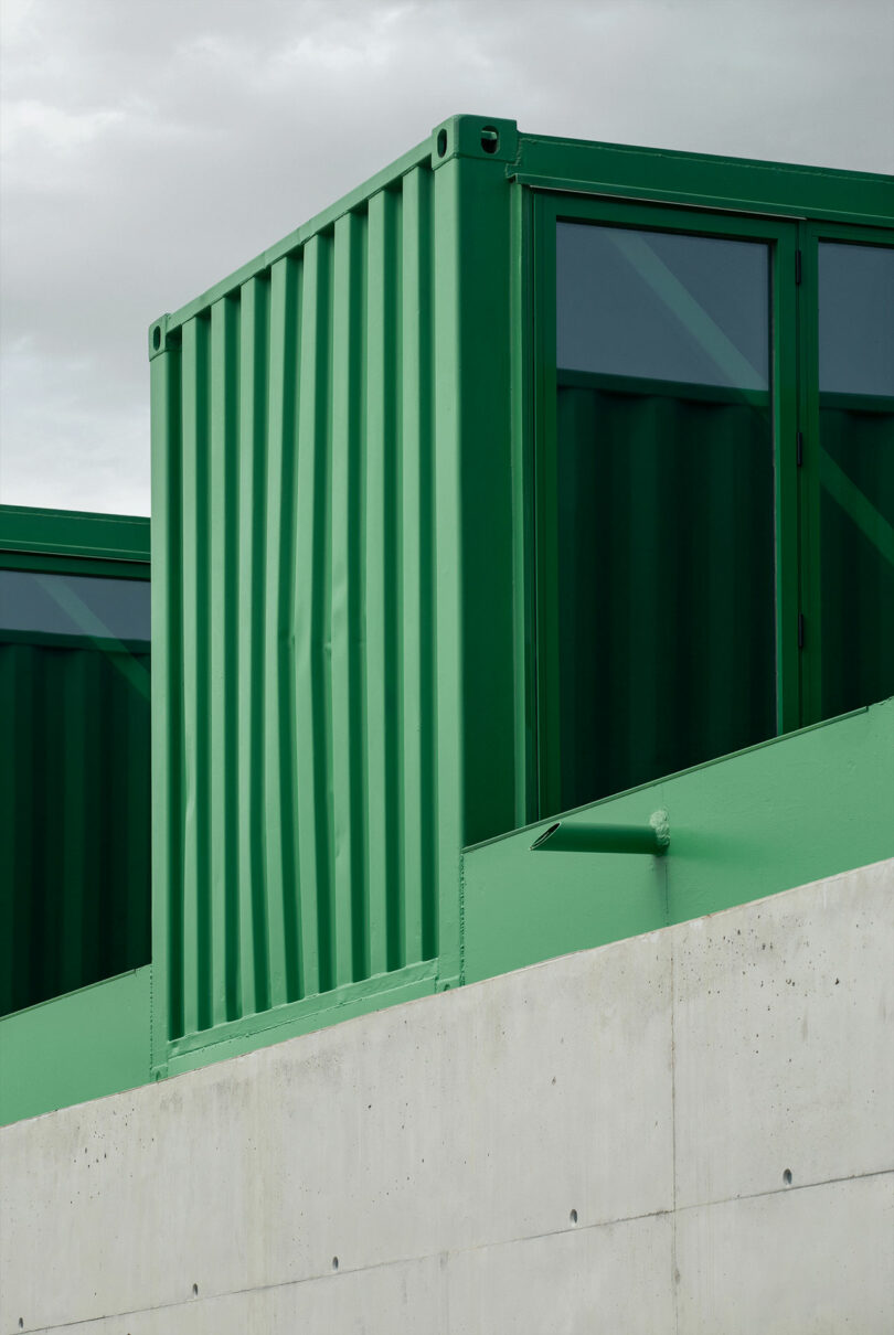 Impepinable Studio’s green shipping container structure is set atop a concrete wall, framed by an overcast sky in the background.