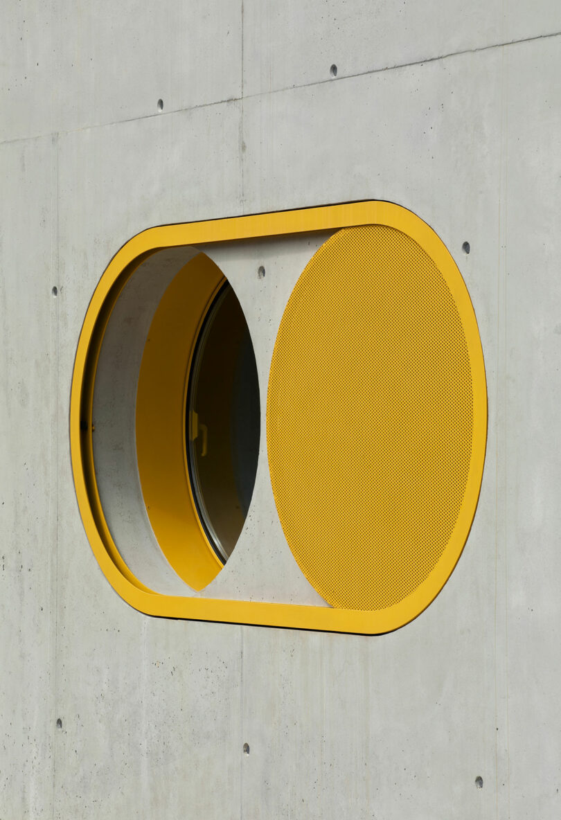 Oval window with a yellow border on a concrete wall, partly covered by a perforated yellow metal screen—an eye-catching detail designed by Impepinable Studio.