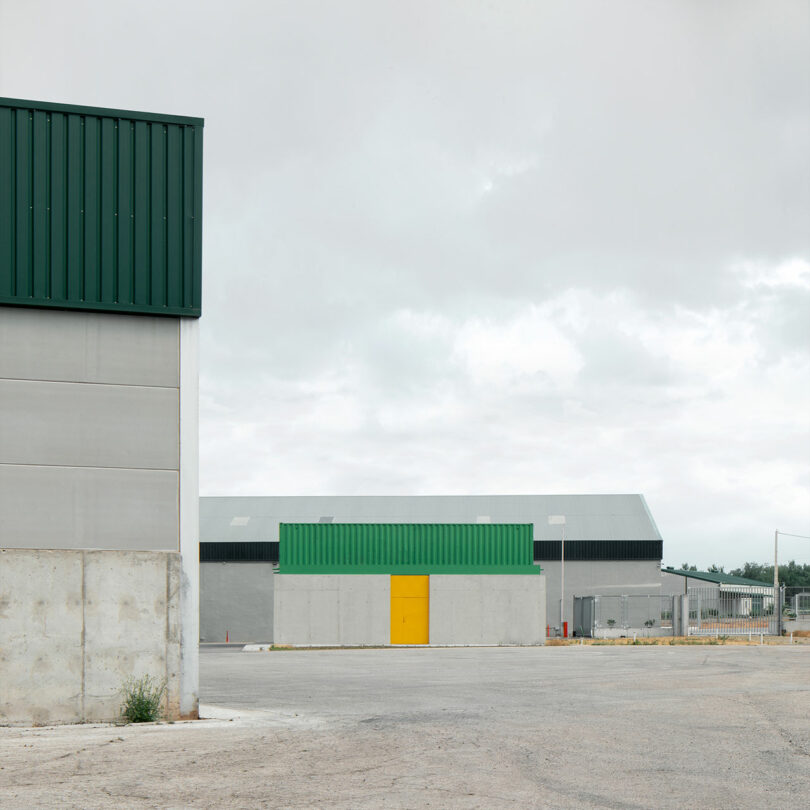 Industrial buildings with green and yellow accents under an overcast sky; empty paved area in the foreground, sparse vegetation around the perimeter—an Impepinable Studio touch gives the scene a distinctive edge.