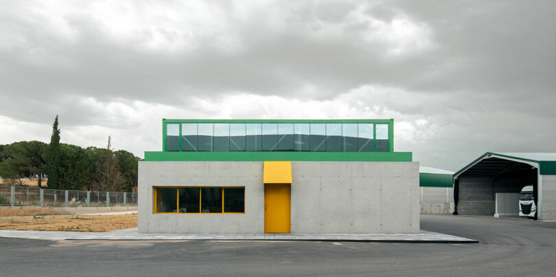 A small, modern concrete building with a green trim, yellow-framed windows, and a yellow door stands in an industrial area under a cloudy sky—home to the creative Impepinable Studio.