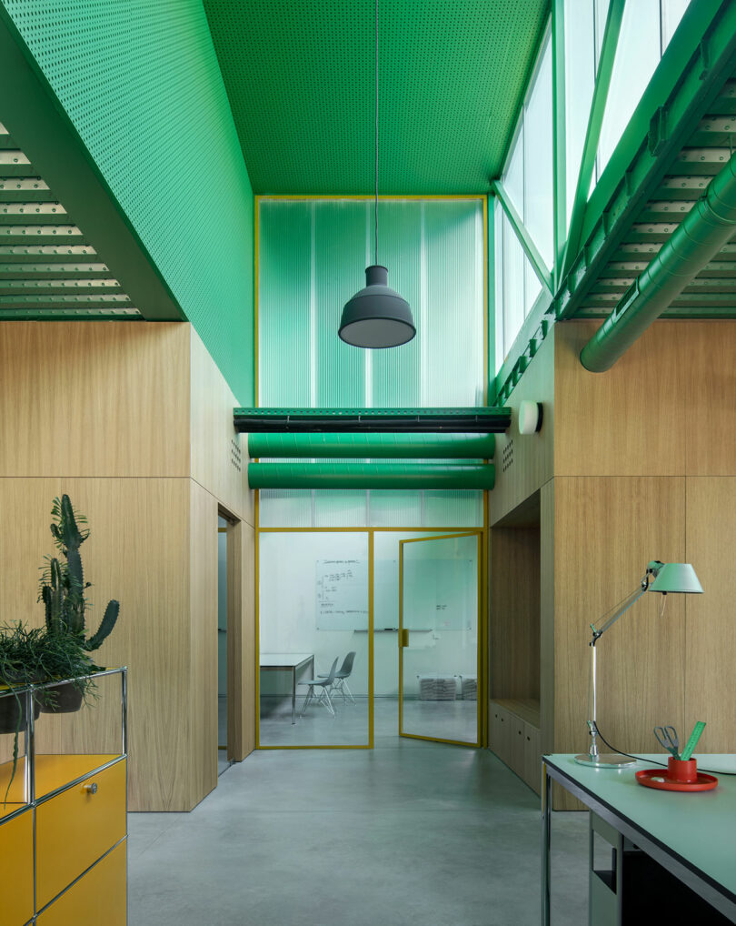 Modern office interior by Impepinable Studio featuring a high green ceiling, wood-paneled walls, glass doors with yellow frames, and minimal furniture including a desk lamp and a cactus plant.