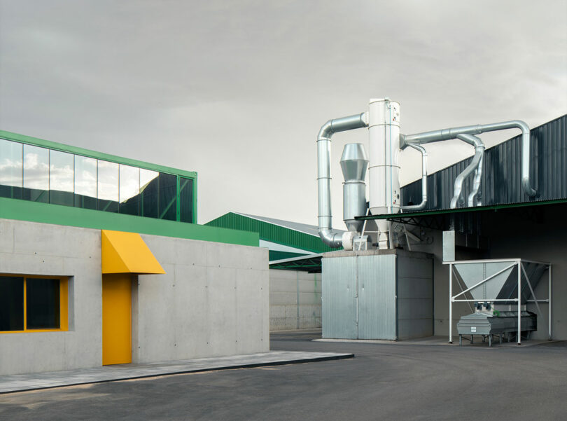 Industrial facility with concrete buildings, a bright yellow door and window, green metal roofs, and large silver ventilation ducts under a cloudy sky—home to the innovative Impepinable Studio.
