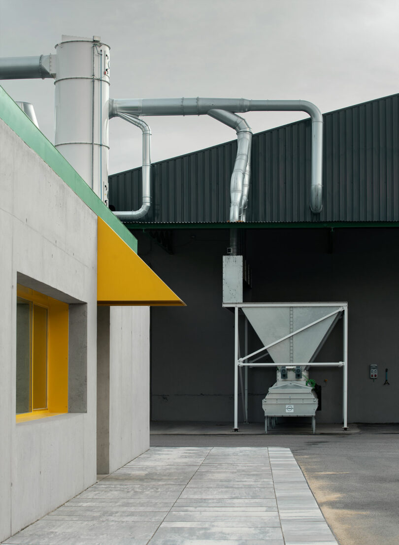 Industrial building exterior with exposed metal ventilation pipes, a yellow window awning, grey walls, and a large metal hopper against the dark wall—an Impepinable Studio aesthetic.