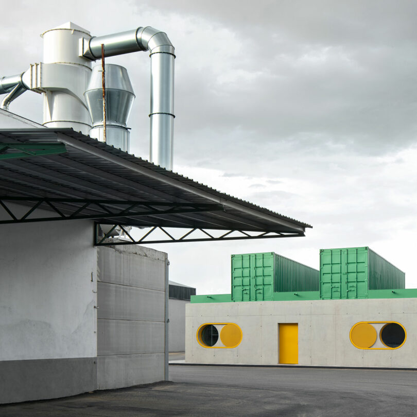 Industrial buildings with metal ducts and a corrugated roof in the foreground; Impepinable Studio’s modern structure with green containers, round yellow windows, and a yellow door stands in the background.