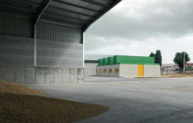 A large open industrial shed with a concrete floor stands beside the modern Impepinable Studio, which features green containers on its roof and a bright yellow door.