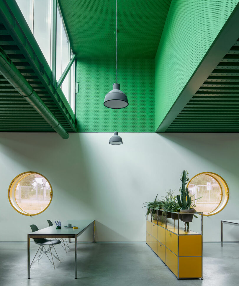 Modern office space by Impepinable Studio with a green ceiling, round yellow windows, yellow storage unit with plants, and a minimalist table with two chairs.