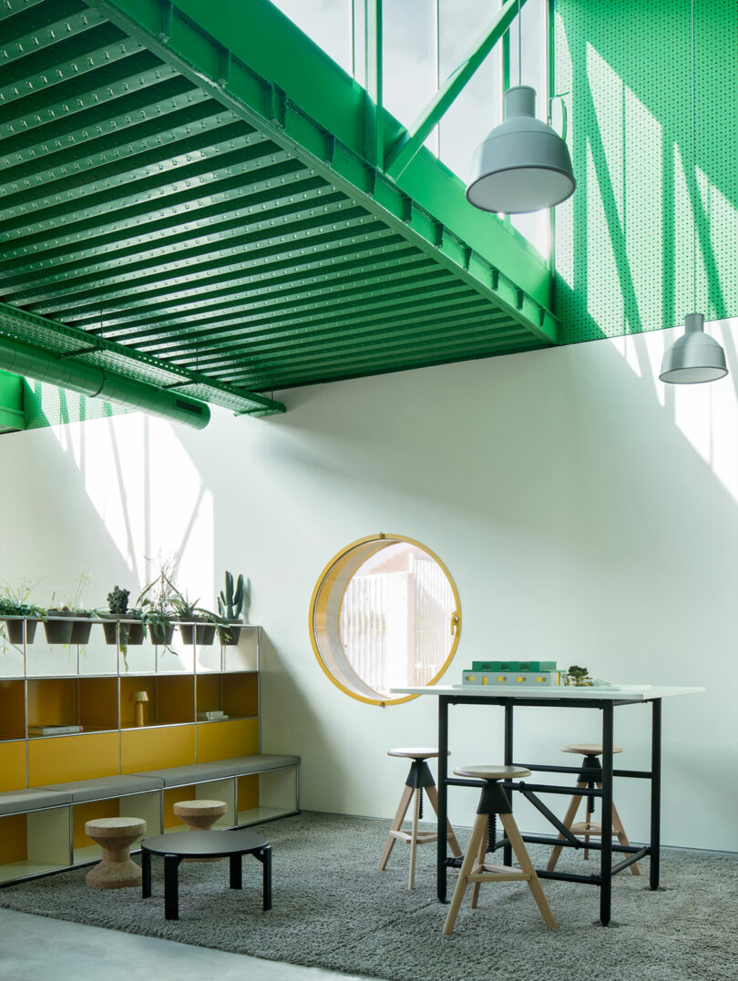 Modern Impepinable Studio workspace with green ceiling beams, yellow shelving, a round window, high table with stools, and plants, all on a gray rug under natural light.