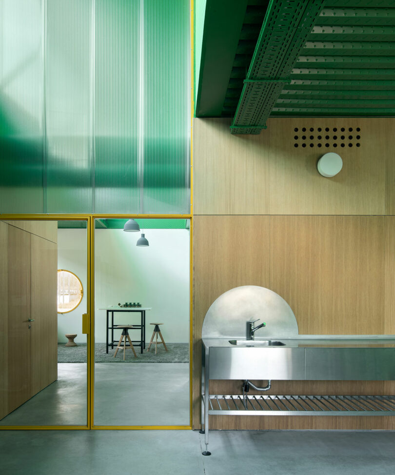 Contemporary kitchen interior by Impepinable Studio features a stainless steel sink, wood paneling, yellow-framed glass door, and green ceiling beams, seamlessly opening into a minimalist adjoining room.