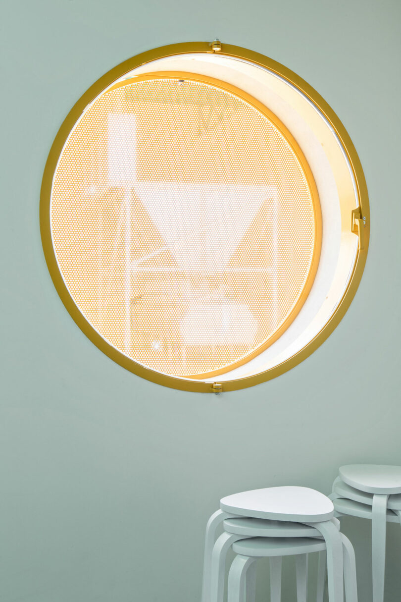 A round yellow-framed window with a perforated screen, designed by Impepinable Studio, is set in a pale green wall above a stack of white stools.