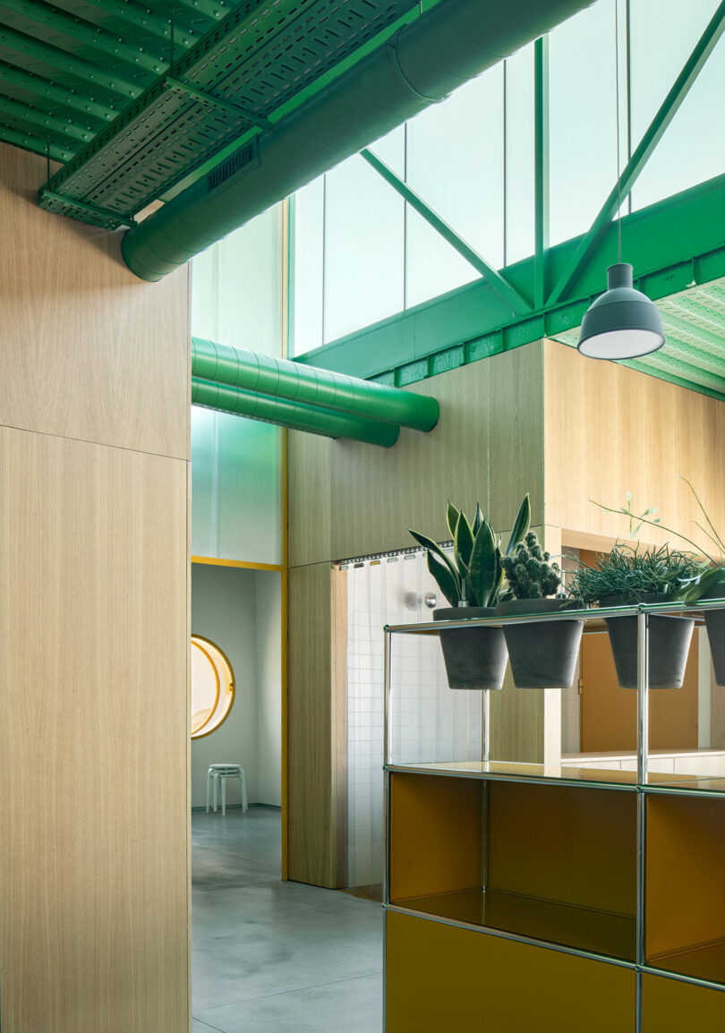 Modern interior with green exposed ducts, wooden wall panels, a round window, and metal shelving with potted plants in the Impepinable Studio. A hanging lamp and natural light beautifully illuminate the space.