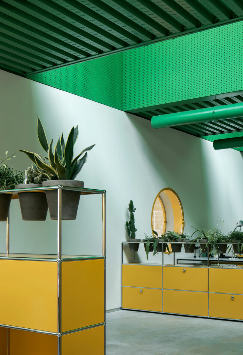 A modern interior by Impepinable Studio with green ceilings and ductwork, yellow modular cabinets, potted plants, and a round mirror on a white wall.