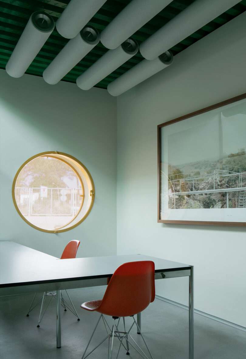 Minimalist office by Impepinable Studio with two orange chairs, a rectangular table, a round porthole window, and a large framed photograph. The green ceiling is highlighted by cylindrical panels.