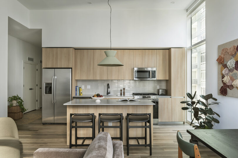 Modern kitchen with light wood cabinets, stainless steel appliances, a central island with three black stools, and a large window providing natural light—perfect for enjoying the Roost Rainey lifestyle.
