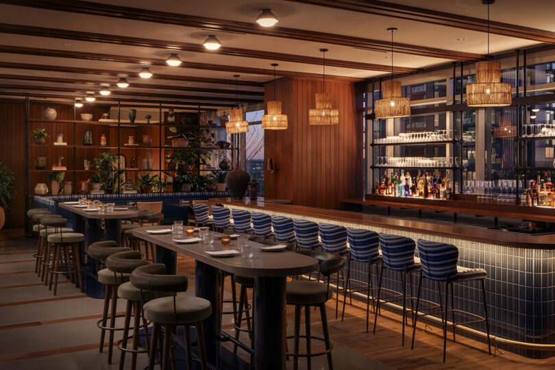 Modern bar and dining area at Roost Rainey with blue cushioned stools, pendant lighting, wood accents, a stocked bar, and plants on shelves. The space is empty and warmly lit.