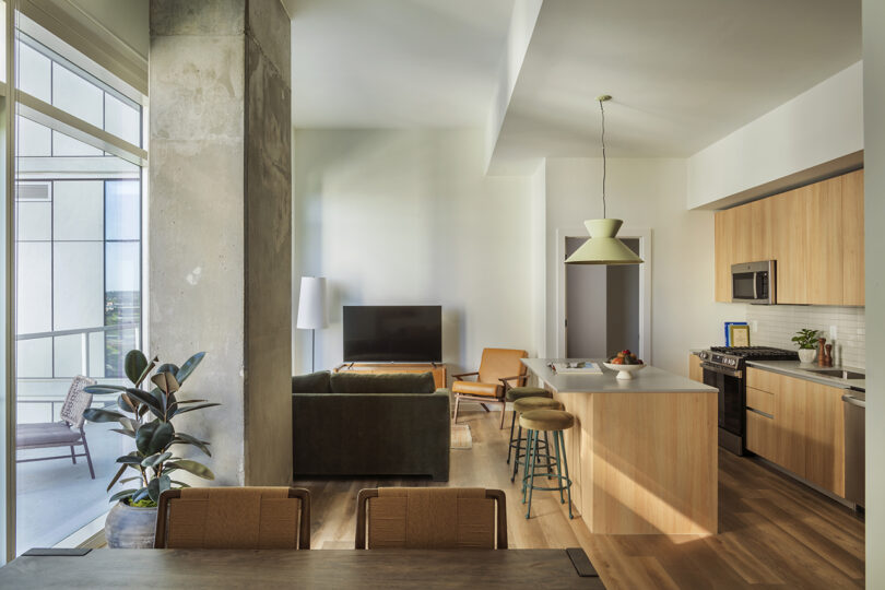 Modern open-plan apartment at Roost Rainey with a kitchen, dining area, and living room. Large window, indoor plants, neutral-toned furniture, and a balcony are visible.