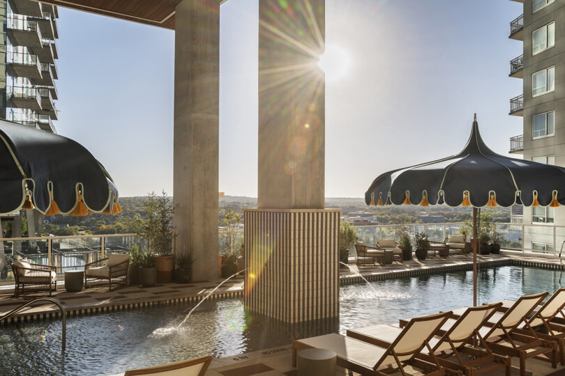 Outdoor pool area at Roost Rainey with lounge chairs, large black umbrellas, and the sun shining behind a tall pillar, surrounded by high-rise buildings.