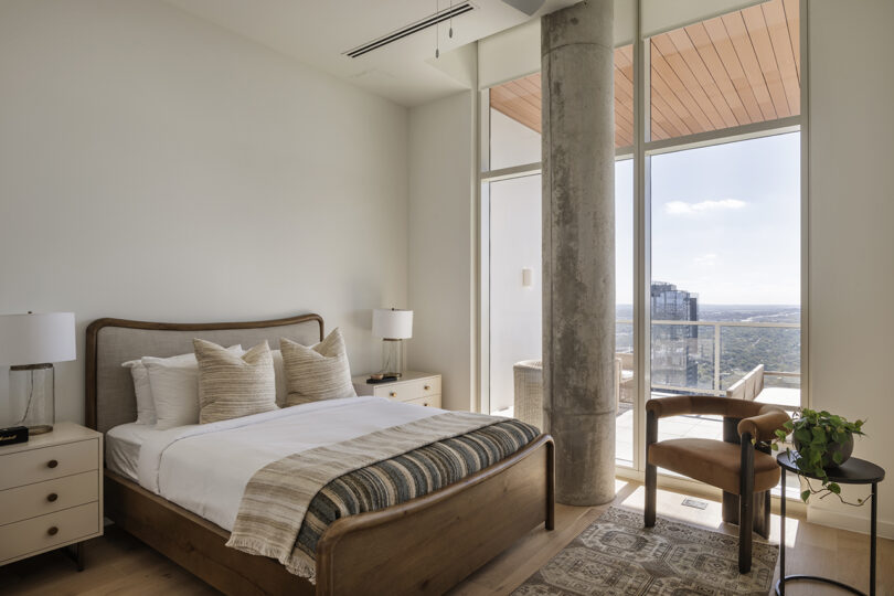 Modern bedroom with a wooden bed, neutral bedding, two nightstands with lamps, an armchair, and a potted plant showcases Roost Rainey style through floor-to-ceiling windows overlooking a balcony with a city view.