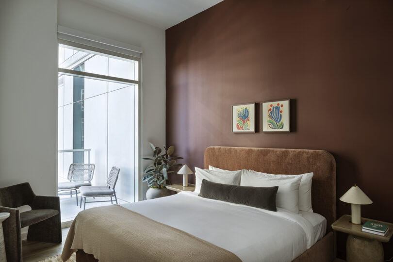A modern Roost Rainey bedroom with a brown accent wall, double bed, two nightstands with lamps, a potted plant, and access to a balcony with two chairs and a small table.