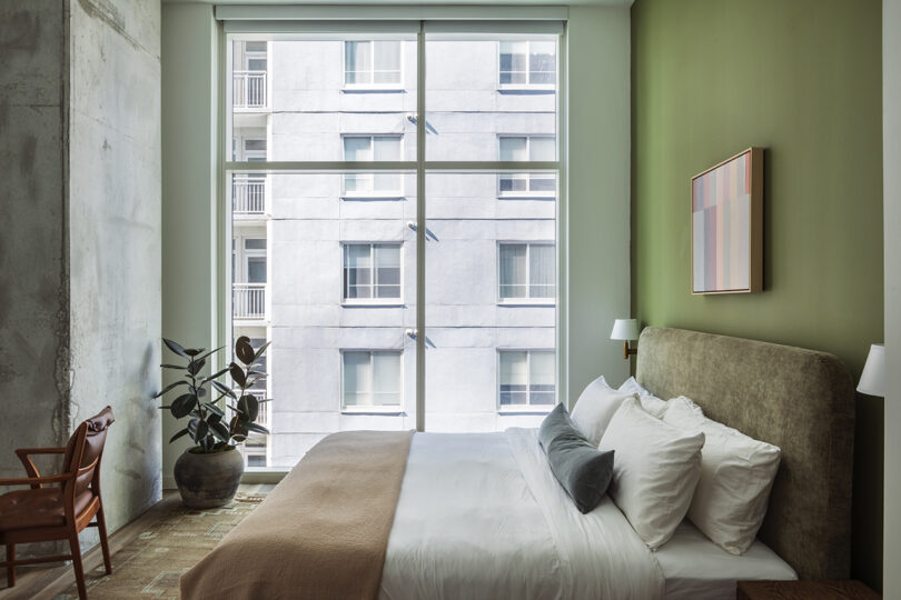 Modern bedroom at Roost Rainey with green accent wall, large window, potted plant, bed with white linens, beige blanket, wall art, and wooden chair. Apartment building visible outside the window.