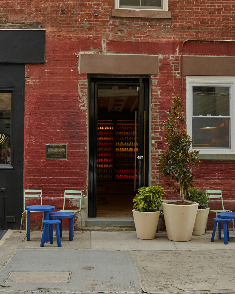 A brick storefront with an open door, three blue tables and chairs, and several large potted plants on the sidewalk. The interior displays shelves with colorful objects.