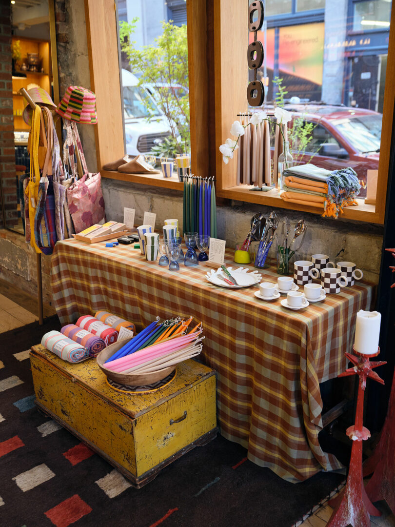 A table covered with a plaid cloth displays umbrellas, teacups, bowls, chopsticks, and various small gift items in a shop with large windows and bags hanging nearby.