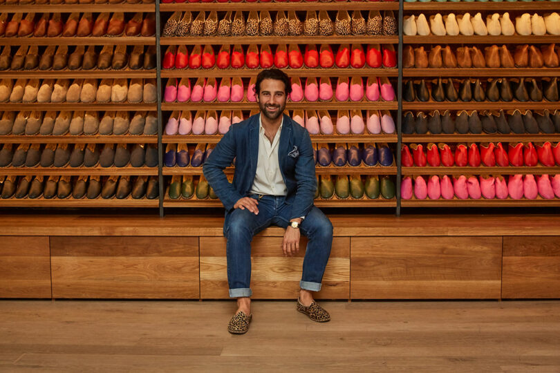 A man in a blue suit sits on a wooden bench in front of shelves displaying many pairs of colorful shoes.