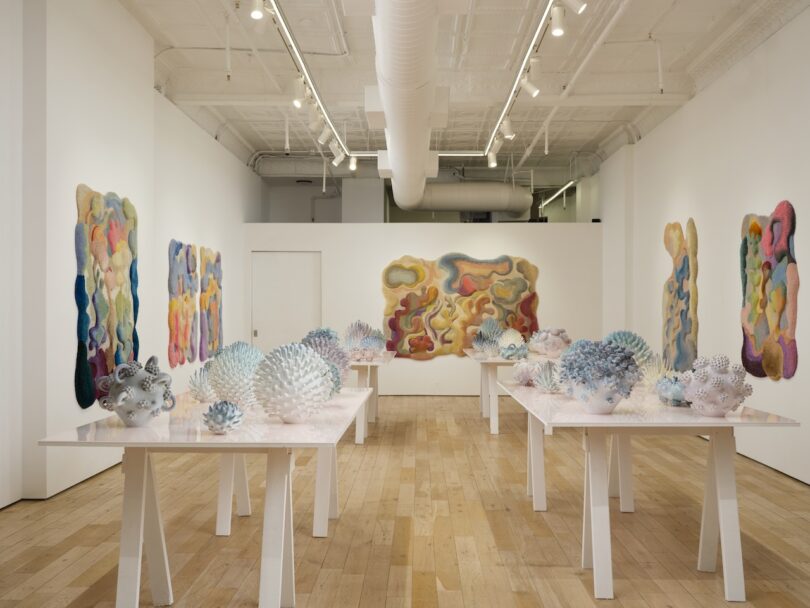 An art gallery displays shimmering, colorful wall hangings and sculptural ceramic forms on white tables arranged in two rows on a wooden floor, creating a scene that feels both abstract and real.