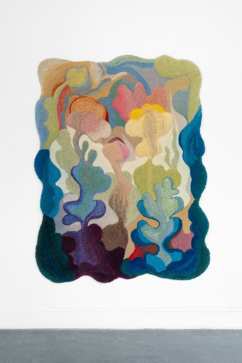 A colorful, abstract textile artwork with shimmering, organic wavy shapes and layered textures is mounted on a white wall above a gray floor, creating a sense of real visual depth.