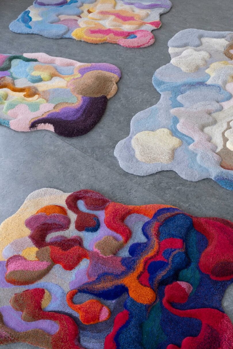 Four irregularly shaped, multicolored rugs with shimmering real abstract patterns are placed on a gray floor.