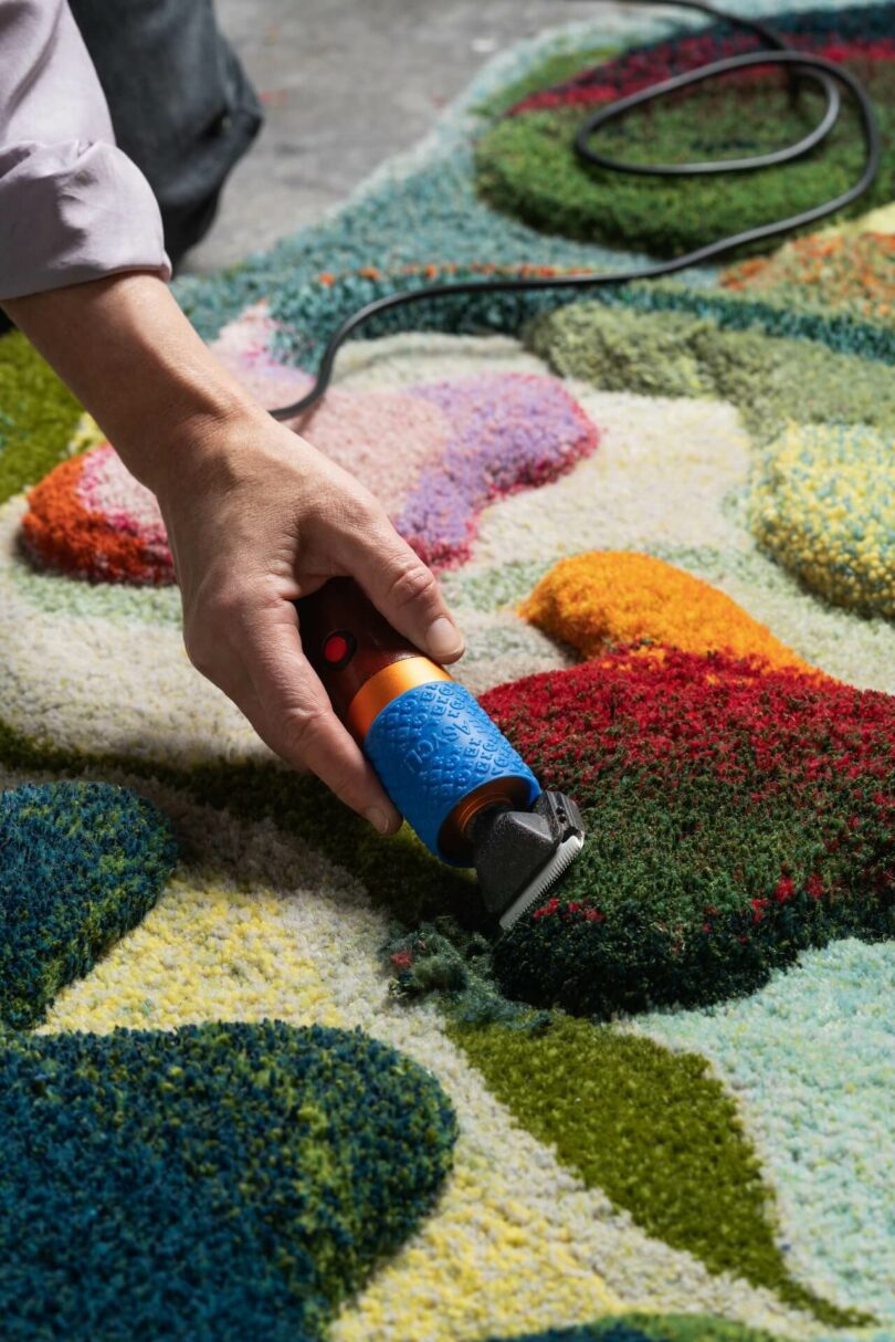 A person uses an electric trimmer to shape a shimmering, colorful rug with various abstract patterns, bringing a touch of shimmering real flair to the textured surface.