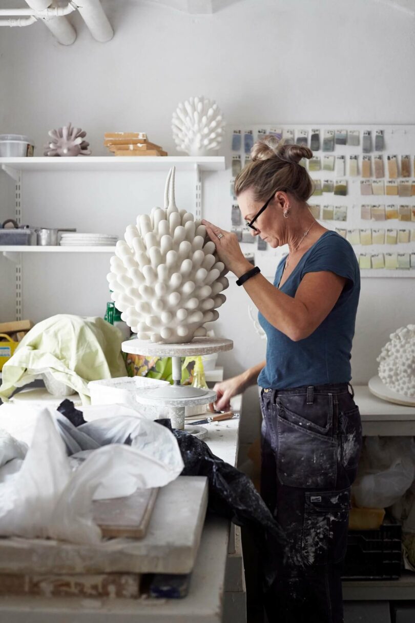 A woman in a studio works on a large, textured ceramic sculpture; shelves with paint samples and art materials are visible in the background, capturing a shimmering real moment of creative focus.
