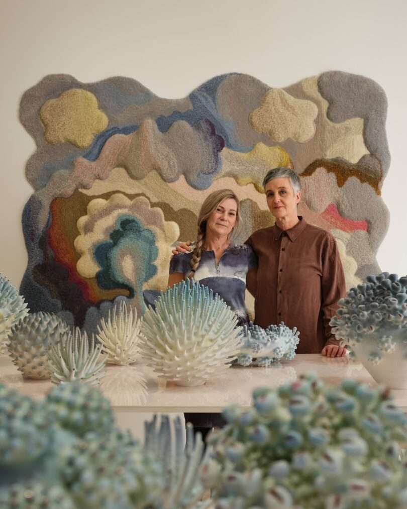 Two people stand together in front of a shimmering, textured wall hanging, surrounded by sculptural ceramic artworks resembling sea urchins and coral, creating a scene that feels both abstract and real.