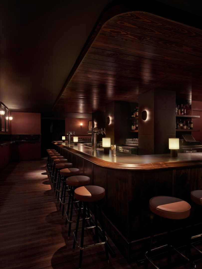 Dimly lit bar interior at Municipal Grand, featuring a long wooden counter, bar stools, modern lamps, and shelves of bottles and glasses behind the bar.