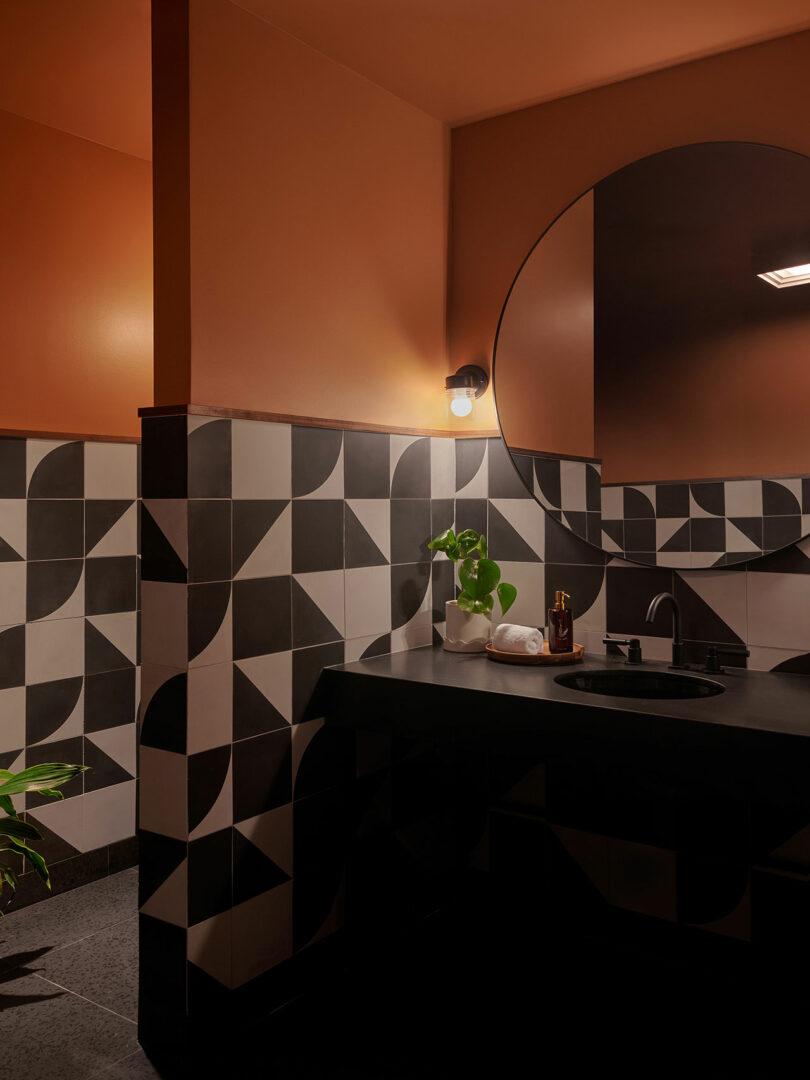 Modern bathroom in a Municipal Grand style, featuring black and white geometric wall tiles, a round mirror, black countertop, and a small plant beside a rolled towel and soap dispenser. Warm lighting glows above.