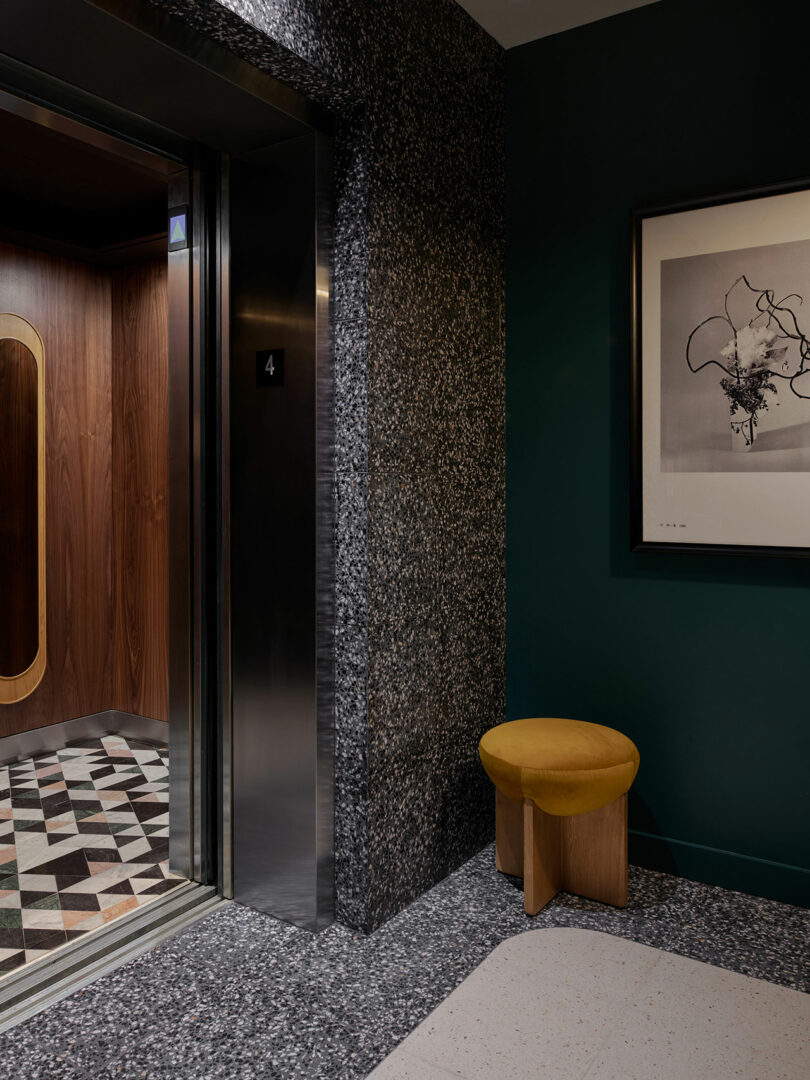 Open elevator with wood interior and geometric tiled floor; nearby, a yellow stool sits against a dark green wall beneath abstract artwork—an inviting nook in the Municipal Grand.