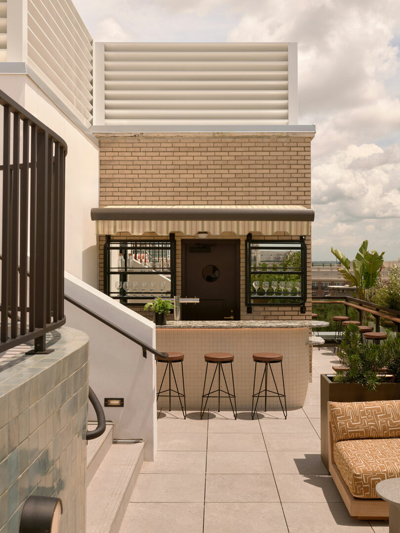 Outdoor rooftop bar at Municipal Grand with four stools, a tiled counter, and a small brick building behind. Cushioned seating and plants add comfort, set against a cloudy sky backdrop.