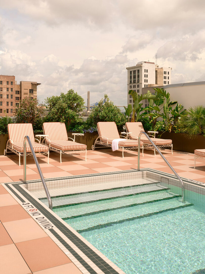Outdoor pool with clear water at Municipal Grand, steps leading in, and striped lounge chairs beside potted plants on a rooftop with city buildings in the background.