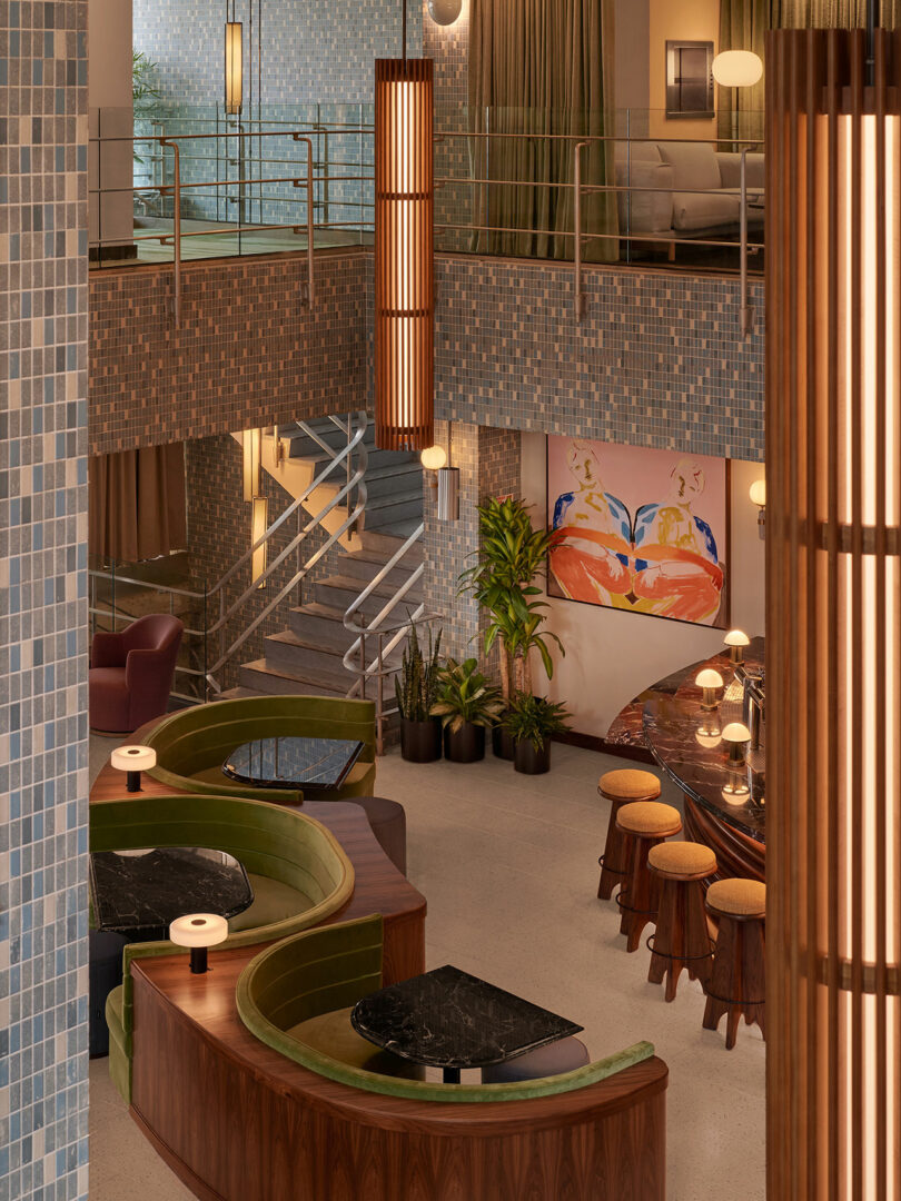 A stylish lounge area at Municipal Grand features curved green booths, marble tables, bar stools, indoor plants, hanging lights, and blue mosaic tile walls. Artwork and a staircase are visible in the background.