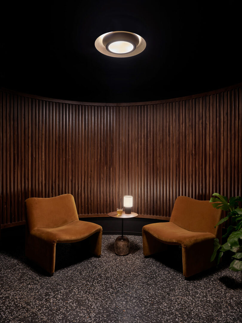 Two brown velvet chairs face each other across a small round table with a lamp and glass, set against a curved wooden slat wall and terrazzo floor beneath a circular ceiling light in the elegant Municipal Grand style.